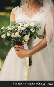 The bride in an elegant wedding dress holds a beautiful bouquet of different flowers and green leaves. Wedding theme.. The bride in an elegant wedding dress holds a beautiful bouquet of different flowers and green leaves. Wedding theme