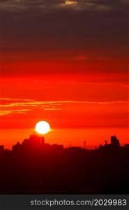 The bright sun in the early morning floods the sky with red light, rising above the silhouettes of the houses of the sleeping city. Vertical image, copy space.. A bright red sun disk rises in the early morning over the silhouettes of city houses. Vertical image.