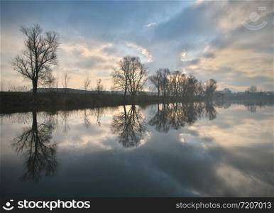 The calm autumn morning landscape with fog and warm sky over the pond surrounded by trees with the beautiful reflections of clouds and trees in the water