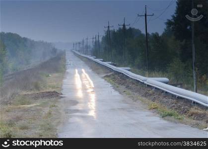 The car runs on wood asphalted road along the pipeline vague cloudy in the morning. The track is laid in the woods, on both sides of the road trees. The road passes through a transmission line.