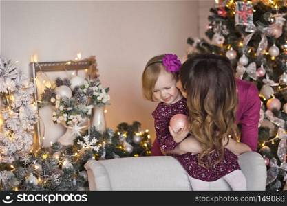 The child with the mother sitting on a white Mat around the Christmas tree.. Portrait of a mother with her daughter play near the fireplace and Christm