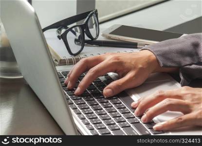 The close-up image of a young woman who is typing on a laptop on a stick in her office and has a notebook, pen and glasses and a lamp placed next to it.