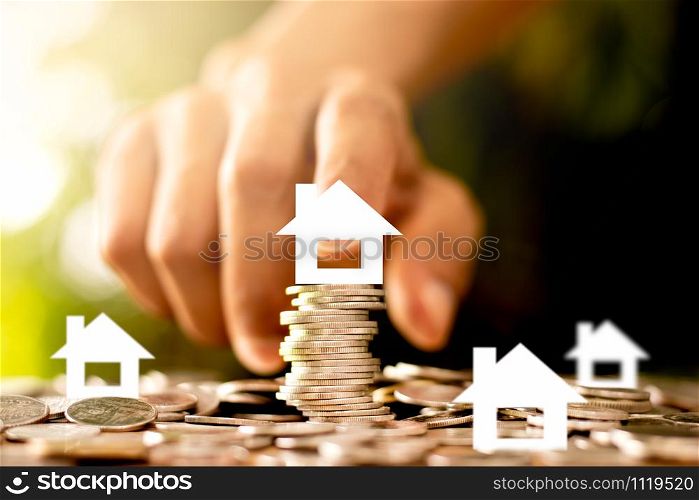 The coins are stacked and there is a white paper house on top. The men&rsquo;s hands are gently touching, the concept of saving money and growing financially.