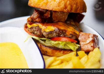 The concept of fast food and takeaway food. A juicy hamburger lies next to french fries on a metal plate along with cheddar cheese sauce in the background. The concept of fast food and takeaway food. A juicy hamburger lies next to french fries on a metal plate along with cheddar cheese sauce in the background.