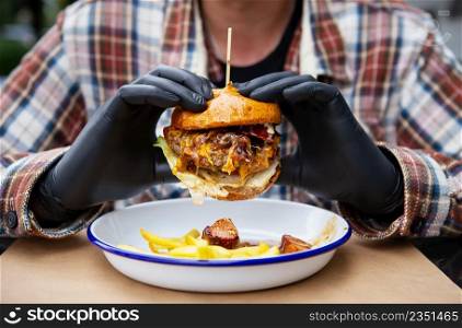 The concept of fast food and takeaway food. A man in black latex gloves holds a juicy hamburger in his hands, lies near french fries on a metal plate along with cheese sauce. The concept of fast food and takeaway food. A man in black latex gloves holds a juicy hamburger in his hands, lies near french fries on a metal plate along with cheese sauce.