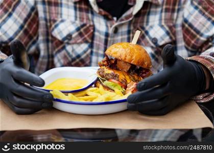 The concept of fast food and takeaway food. A man in black latex gloves is ready to eat a juicy hamburger that lies near the french fries on a metal plate along with cheese sauce. The concept of fast food and takeaway food. A man in black latex gloves is ready to eat a juicy hamburger that lies near the french fries on a metal plate along with cheese sauce.