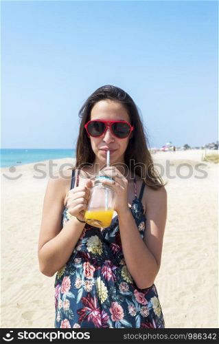 The concept of leisure and travel. A young tanned girl came to the resort, wears a fashionable dress and sunglasses, holds a delicious juice drink in her hand.