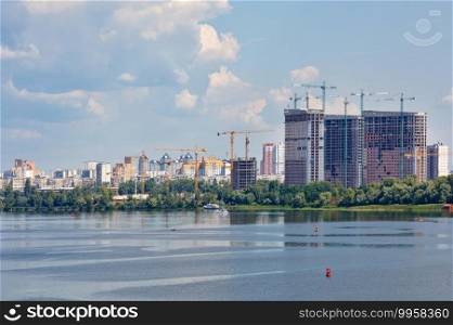 The construction of modern residential buildings rises above the wide surface of the river against the backdrop of the cityscape, copy space.. Cityscape, new residential buildings are being built on the banks of the Dnipro River.