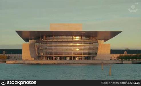 The Copenhagen Opera House at the shore of the harbour at sunset. Modern building with glassy facade and golden sunlight reflecting in it