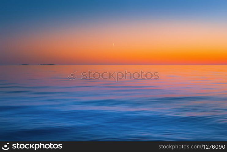 The crescent moon in a clear sky after red yellow sunset reflected in the flowing water surface. Abstract motion blurred seascape background in orange blue colors with copy space. Russia, Lake Onega.