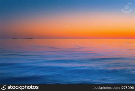 The crescent moon in a clear sky after red yellow sunset reflected in the flowing water surface. Abstract motion blurred seascape background in orange blue colors with copy space. Russia, Lake Onega.