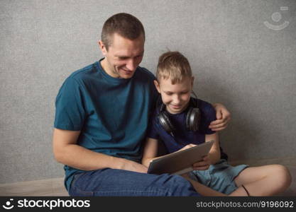 The cute boy took off his headphones and sits on the floor with his dad and looks at the tablet. Dad hugs his son sitting on the floor with big headphones, playing in the gadget
