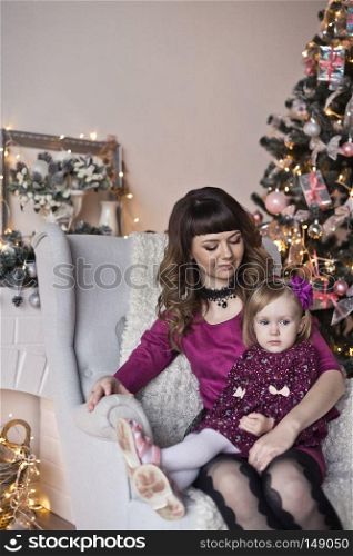 The daughter and mother sitting in a chair near the Christmas tree.. Portrait of a child and its mother sitting on a chair near the Christmas tree