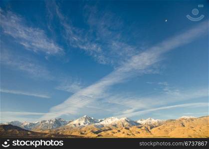 The Eastern Sierra mountain range showing big sky, clouds and the moon in daylight.
