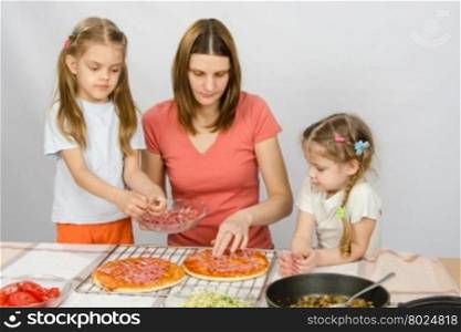 The eldest daughter helps her mother cook a pizza, and the youngest is watching them