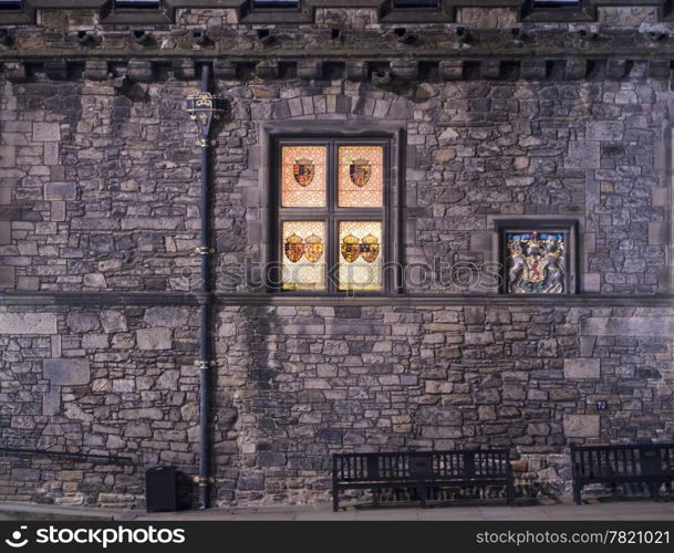 The exterior wall of the Great Hall of Edinburgh Castle at night. The stained glass windows with the emblems of past kings of Scotland are glowing in the dark next to the symbol of royalty.