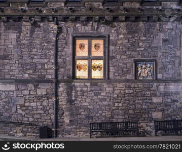 The exterior wall of the Great Hall of Edinburgh Castle at night. The stained glass windows with the emblems of past kings of Scotland are glowing in the dark next to the symbol of royalty.