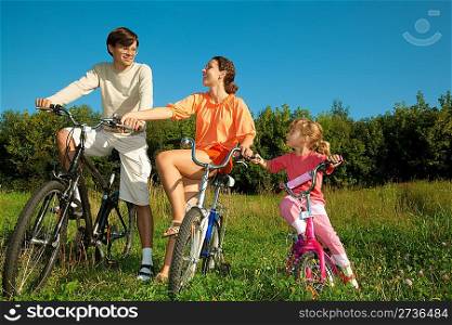 The father, mum and daughter on bicycles in park. To keep the friend for the friend hands.