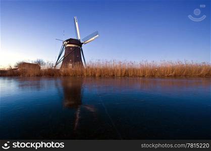 The first rays of sunlight hitting the reed waterfront and the windmill, perched on the side of an ice covered canal on a cold early winter morning