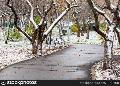 the first snow on bench and lawn and wet path in city park on autumn day