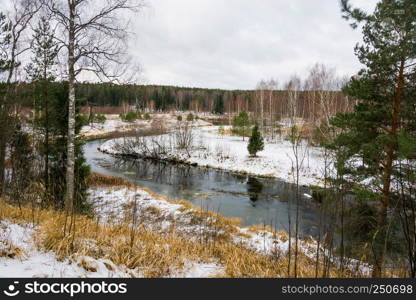 The first white snow fell on the yellow dry grass on the bank of a small river on an autumn cloudy day.