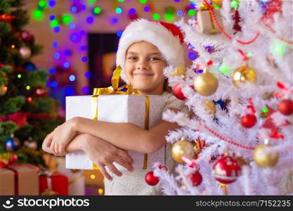 The girl at the Christmas tree holds a big gift in her hands