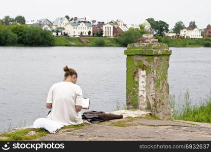 The girl draws a picture sitting on the bank of the river near the old post