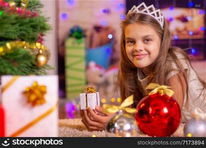 The girl is lying at the Christmas tree and holding a small gift