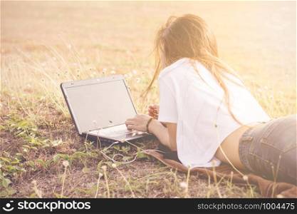 The girl is lying down using a laptop in the park with the sun in the morning.