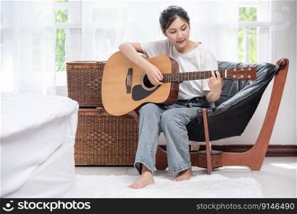The girl is sitting and playing the guitar on the chair.