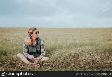 The girl is sitting cross-legged in the field, she is wearing dark glasses and is looking to the side into the distance. Portrait of girl in glasses sitting cross legged in the field. Relaxed teen girl sitting in the field looking to the side. Concept of young woman sitting and relaxing in the field
