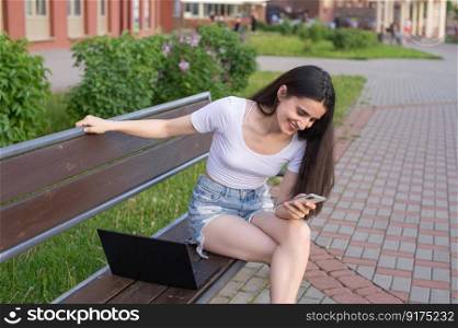 The girl is sitting on a bench, laughing and looking at the phone, there is a laptop nearby.. The girl is sitting on a bench, laughing and looking at the phone, there is a laptop nearby