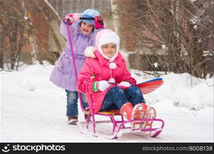 The girl rolls the other girl on a sled in the yard. Two girls girlfriends ride each other on a sled in the snowy winter weather