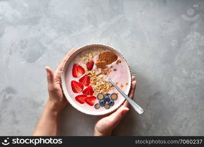 The girl&rsquo;s hands take a plate with natural yoghurt, berries, granola and almonds, top view. A healthy and light breakfast or snack.. Female hands hold a plate with a spoon and apetite yogurt with blueberries, halves of strawberries and almonds on a gray concrete background