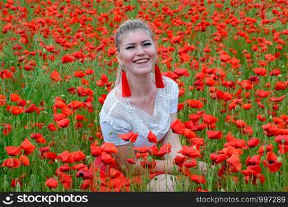 The girl sat in the middle of a poppy field. Blonde young woman in a red skirt and white shirt, red earrings is in the middle of a poppy field.. girl sat in the middle of a poppy field.
