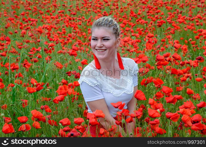 The girl sat in the middle of a poppy field. Blonde young woman in a red skirt and white shirt, red earrings is in the middle of a poppy field.. girl sat in the middle of a poppy field.