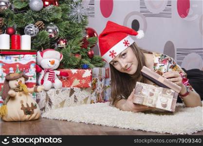 The girl sitting at the Christmas tree. In her hands a big box with a present. Girl opens the box looks in her