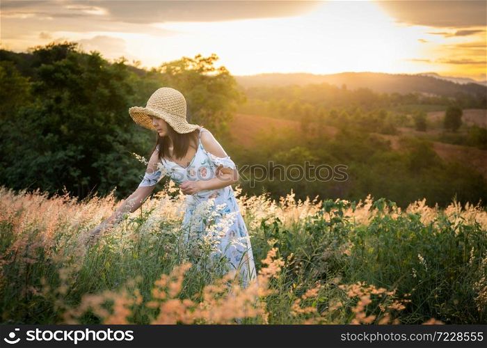 The girl wearing a hat, wearing a white dress, standing in the middle of the grass with beautiful white flowers with a relaxed and happy mood on mountain in sun set time.