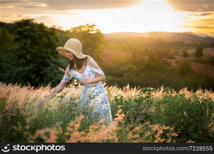 The girl wearing a hat, wearing a white dress, standing in the middle of the grass with beautiful white flowers with a relaxed and happy mood on mountain in sun set time.