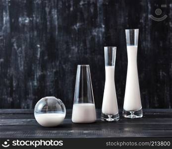 The glasses with dairy product milk standing on a black wooden table on a black background with copy space. Natural organic dairy product.. Preservation technology of natural organic dairy product milk in a glass bottles different shapes on a black wooden table.