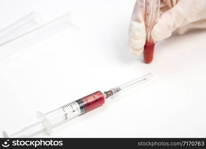 the gloved hand of a laboratory assistant holds a test tube with a blood sample on a white background. also in the photo is a syringe with a blood sample