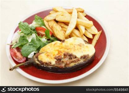 "The Greek dish of aubergines stuffed with minced beef, onion and tomatoes, topped with bechamel sauce and cheese, served with a salad of rocket, lettuca and tomato, and french fried potato chips, viewed from above. The dish is called melitzanes papoutsakia, or "aubergine little-shoes""