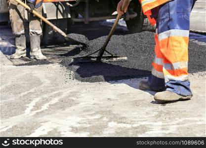 The group of road workers renews a section of the road with fresh hot asphalt using a spade and wooden level and evenly distributes it over the surface.. The working team evenly distributes hot asphalt with a shovel and wooden level manually over the repaired road section.