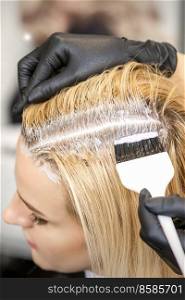 The hairdresser dyeing blonde hair roots with a brush for a young woman in a hair salon. Hairdresser dyeing blonde hair roots