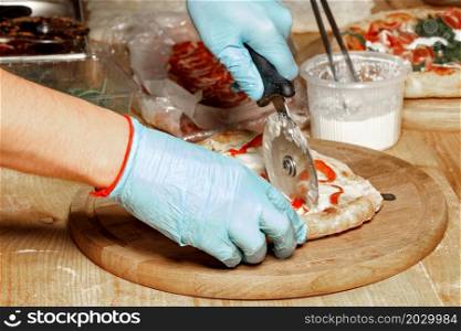 The hands of a chef, a caucasian man in blue latex gloves, are cutting the cooked pizza on a wooden kitchen board. Close-up.. The chef?s hands in blue latex gloves cut the cooked pizza with a round knife.