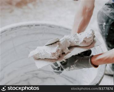 The hands of a young Caucasian male plasterer in construction gloves apply fresh putty with one spatula to another, picking up from a black large basin in the room of his apartment, close-up side view. The concept of wall puttying, home renovation, plastering services, construction work.. The hands of a Caucasian male plasterer i apply fresh putty with one spatula to another