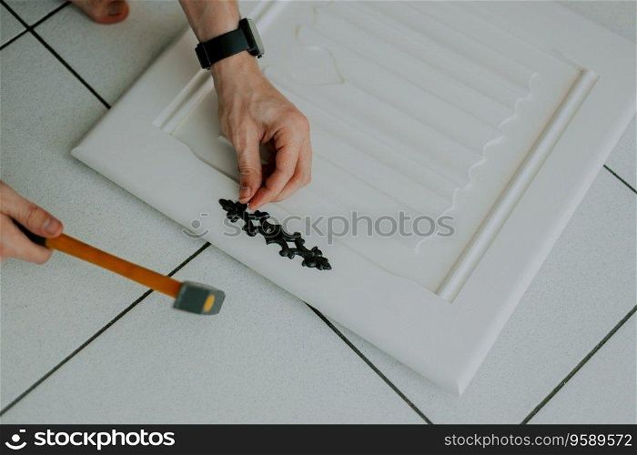 The hands of a young Caucasian unrecognizable man hammers a nail, installing an antique carved black lock on a white cabinet door, close-up top view.. A young guy is repairing a retro lock on a locker door.