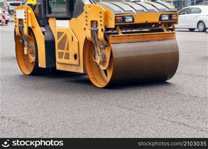 The heavy metal cylinders of the yellow road vibratory roller compact the fresh asphalt on the city street. Copy space.. A road roller compacts fresh asphalt on the roadway on a city street.