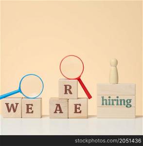the inscription we are hiring and a wooden figurine on the podium with a plastic magnifying glass on a beige background. Employee search concept, open vacancies in the company