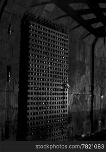 The inside castle gate to Edinburgh Castle at night shows a little bit of light reflecting off the metal studs in the door and shadows from the portcullis. In black and white.